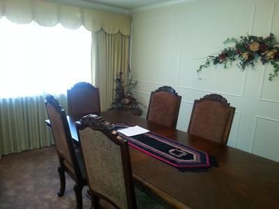 Dining room with a wooden table, six chairs, a runner, and bright curtains by a large window.
