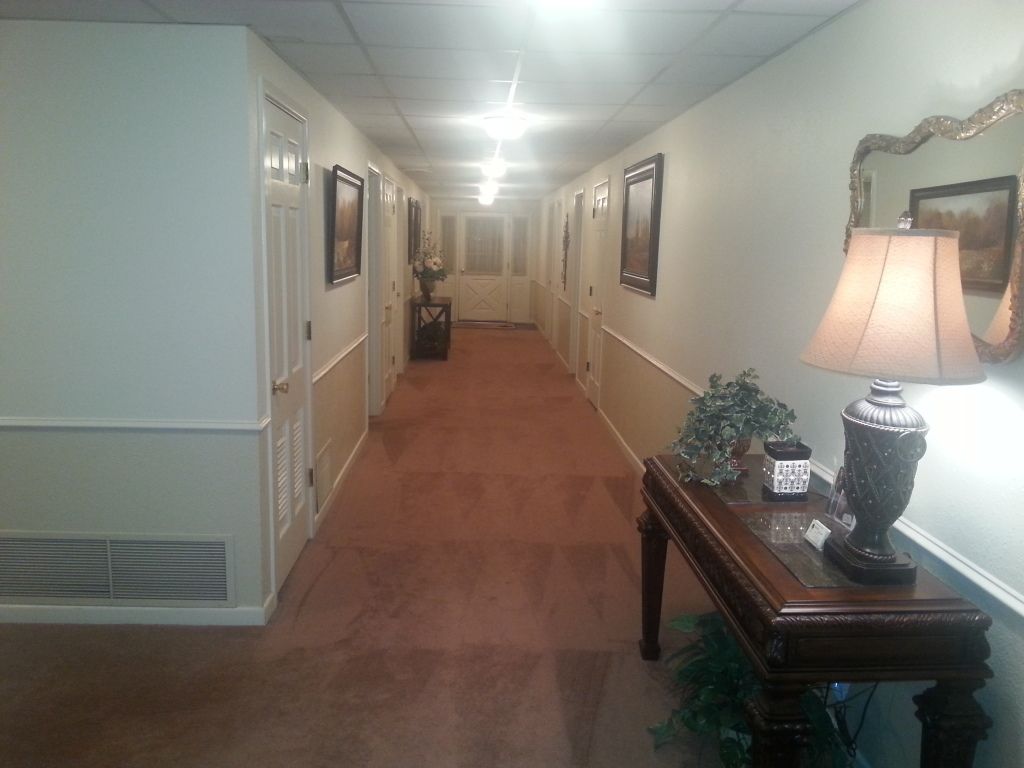 Long hotel hallway with carpet, framed art, and a lamp on a table in the foreground
