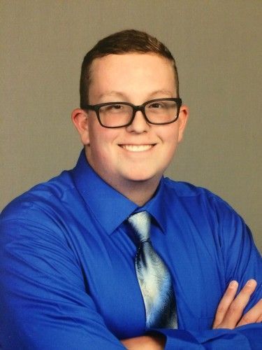 Smiling person in a blue shirt and tie, wearing glasses, with arms crossed against a plain background