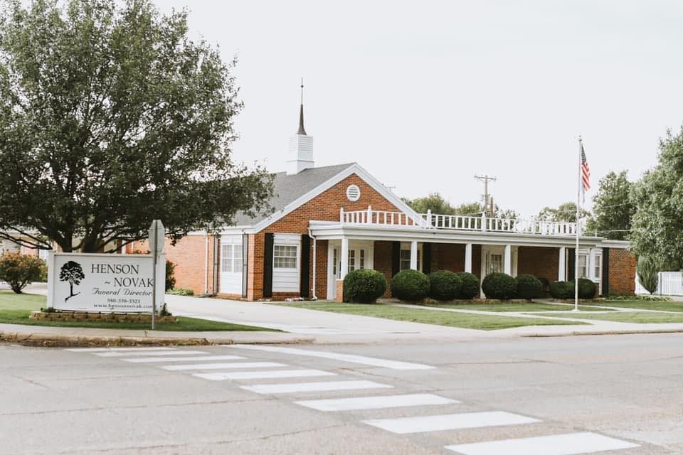 Front entrance of a brick building with white columns, landscaping, and a roadside sign under cloudy skies