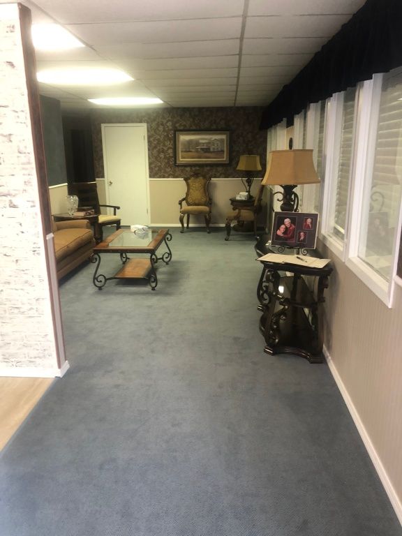 Empty office lounge with gray carpet, sofas, coffee table, and windows along the right wall.