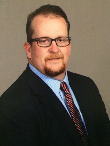 Portrait of a man in a dark suit, blue shirt, and striped tie, wearing glasses, against a neutral background