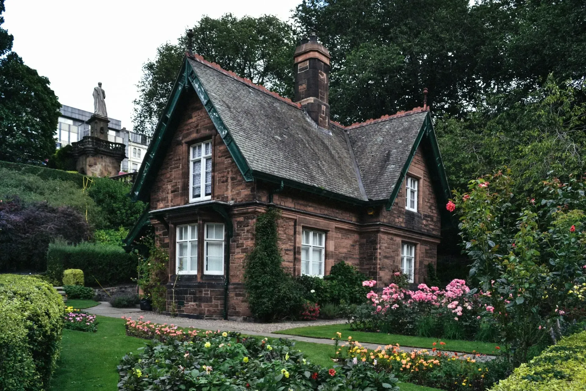 A small brick house with a green roof is surrounded by trees and flowers.