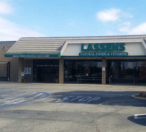 A Lassens Natural Foods & Vitamins store exterior under a blue sky, with a stop sign painted on the asphalt parking lot.