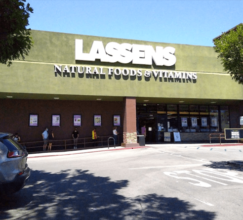 A Lassens Natural Foods & Vitamins store with a green facade, brown brick exterior, and a few people waiting in line.