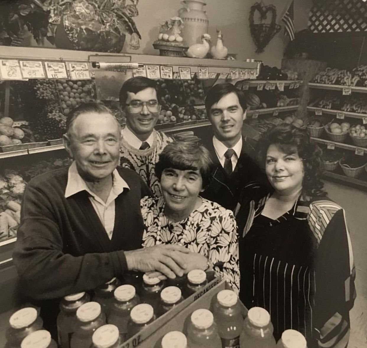 People pose at a market with jars of liquid. Fruits and shelves visible.