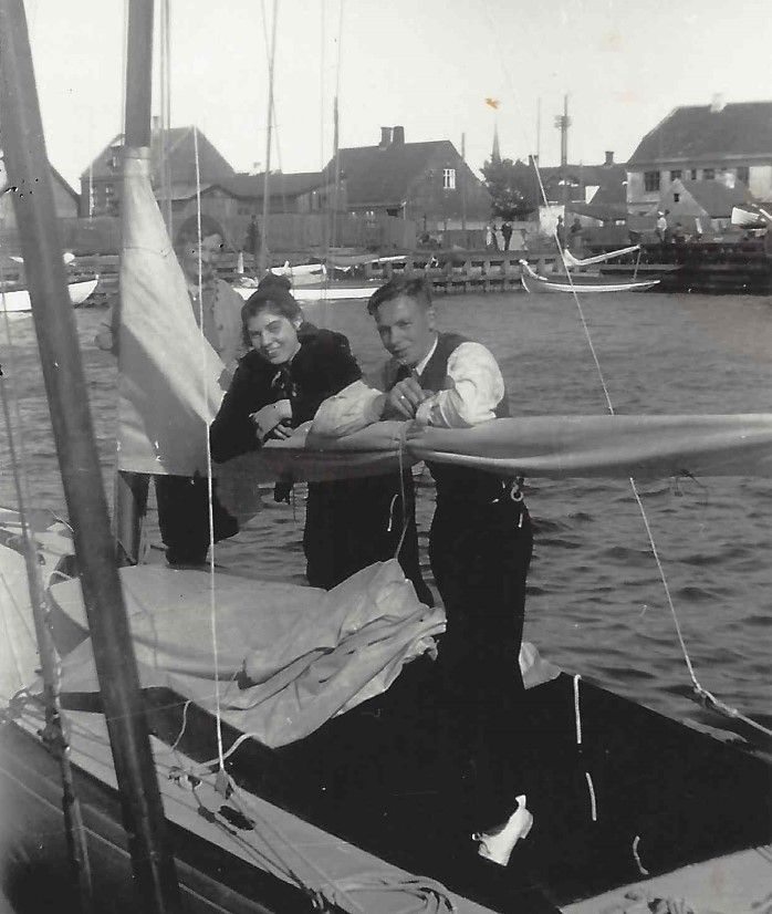 Two people on a sailboat looking at the camera. They are leaning against a rail with a town in the background.