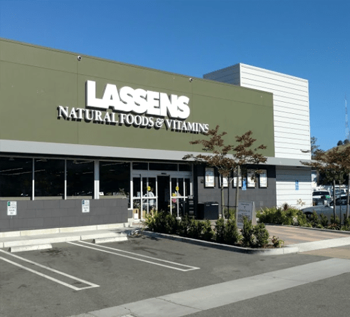 A Lassens Natural Foods & Vitamins store exterior with a green facade, glass entrance, and parking spots under a blue sky.