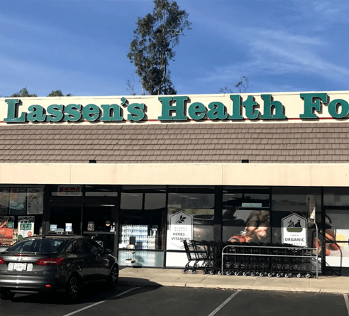 Lassen's Health Foods storefront under a blue sky, featuring a dark grey car parked in front and a row of shopping carts.