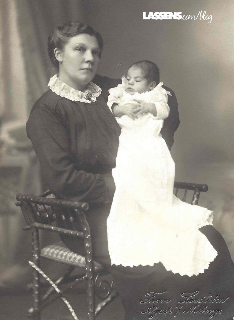 Woman in dark dress holds a baby in a white gown, seated near a chair. Studio portrait.