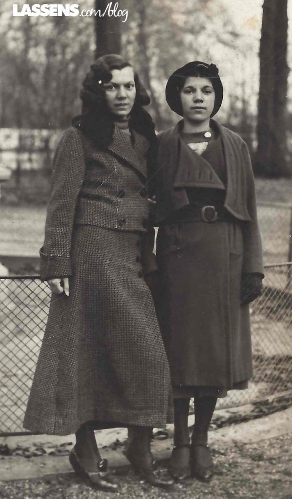 Two women stand side-by-side in a park, wearing long coats and mid-calf dresses, captured in a vintage sepia photograph.
