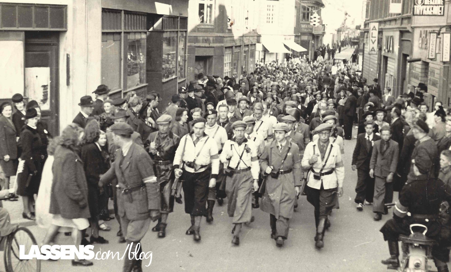 Parade of uniformed people marching down a street lined with onlookers; buildings in the background.