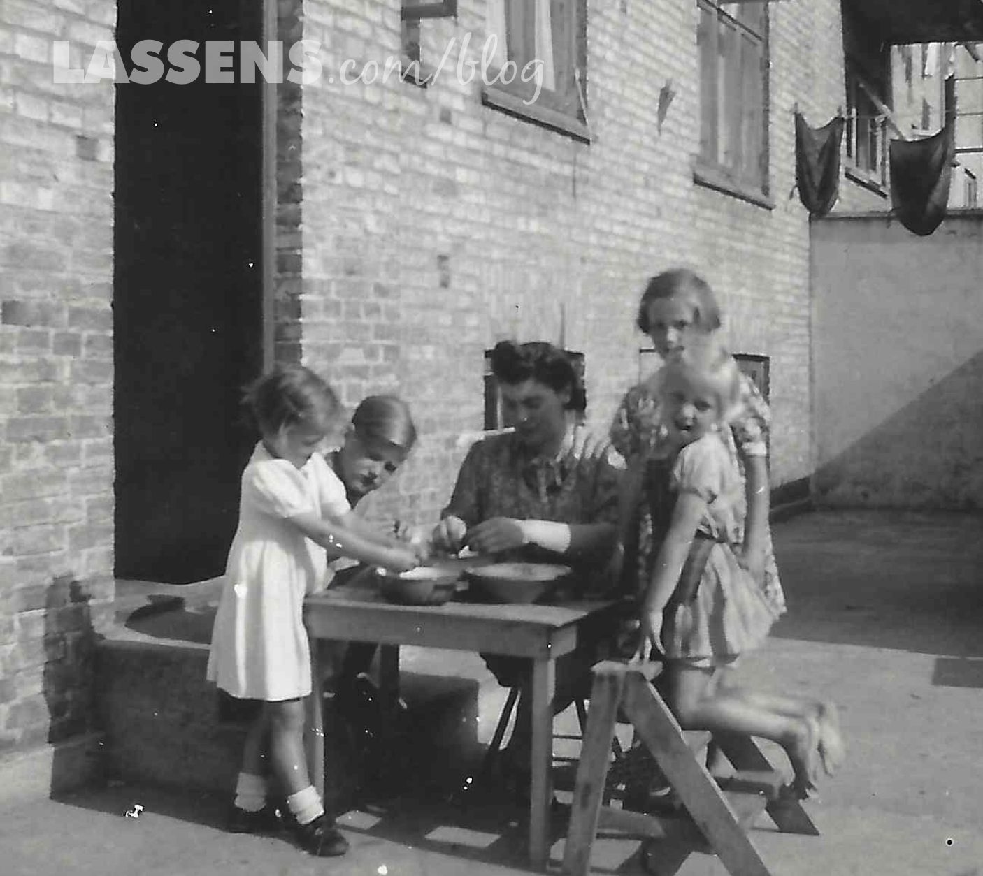 Children and woman sit at table on patio; preparing food. Brick building in background.