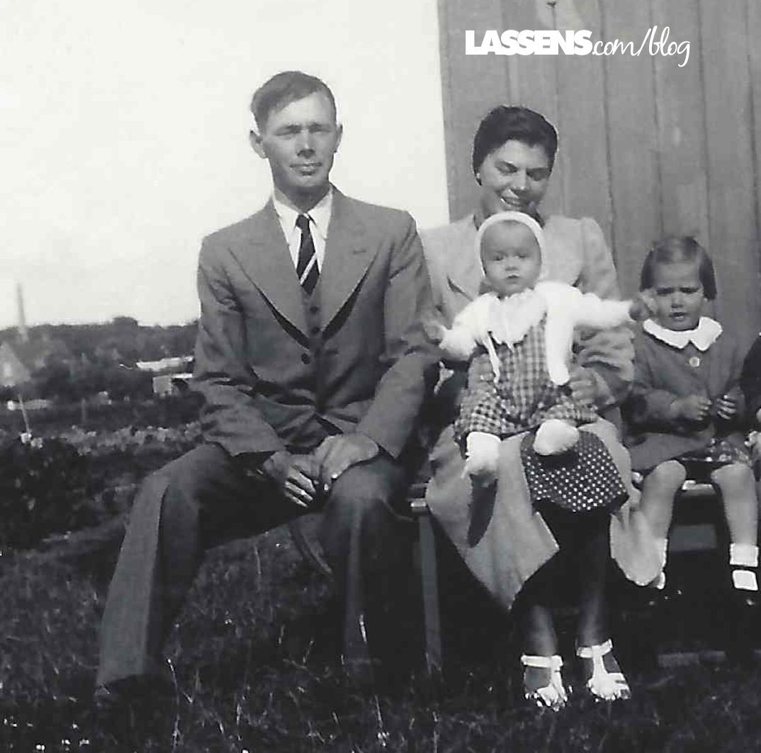 Family of four seated outside. Man in suit, woman holding baby, girl next to her. Black and white photo.