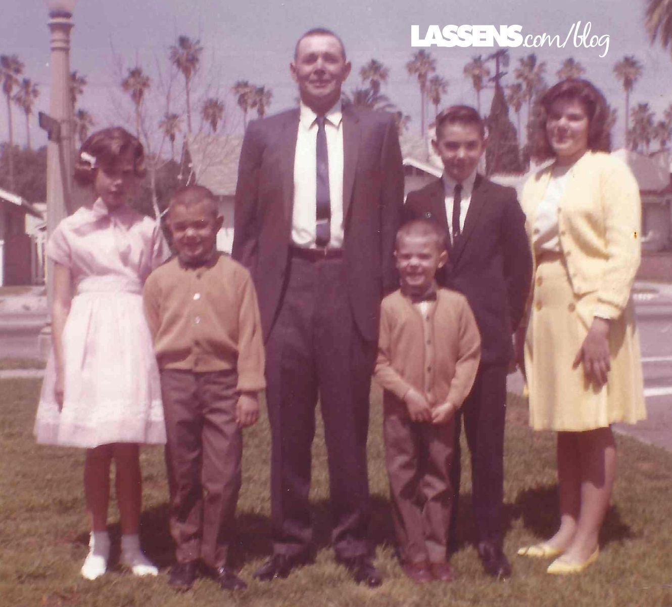 Family of six poses outdoors: man in suit, woman in yellow dress, three boys, and a girl in pink.