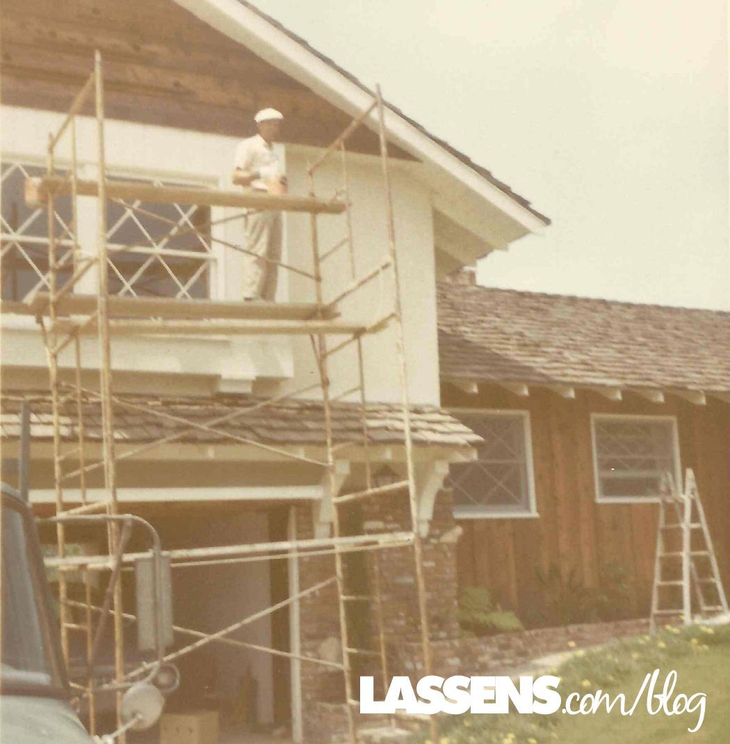 Man on scaffolding painting a two-story house. Wooden shingle roof, windows with geometric grids, and a ladder.