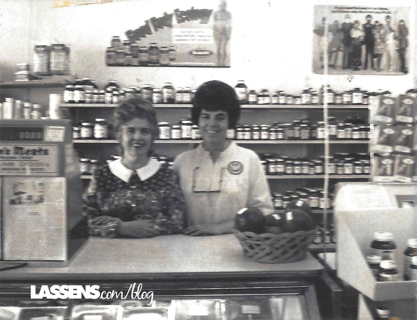 Two women stand behind a store counter filled with jars. Shelves behind are also full of jars. Black and white photo.