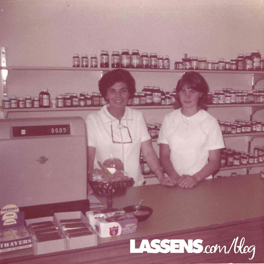 Two women stand behind a counter in a store with shelves of products.