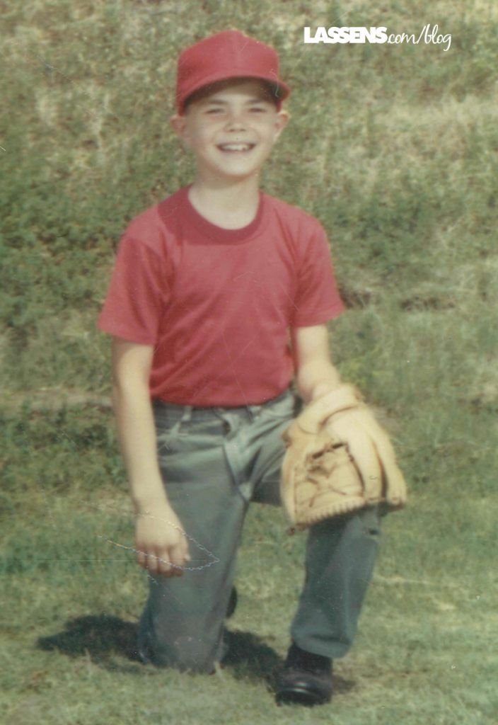 Boy kneeling in a grassy field, wearing a red cap and shirt, denim pants, and a baseball glove.