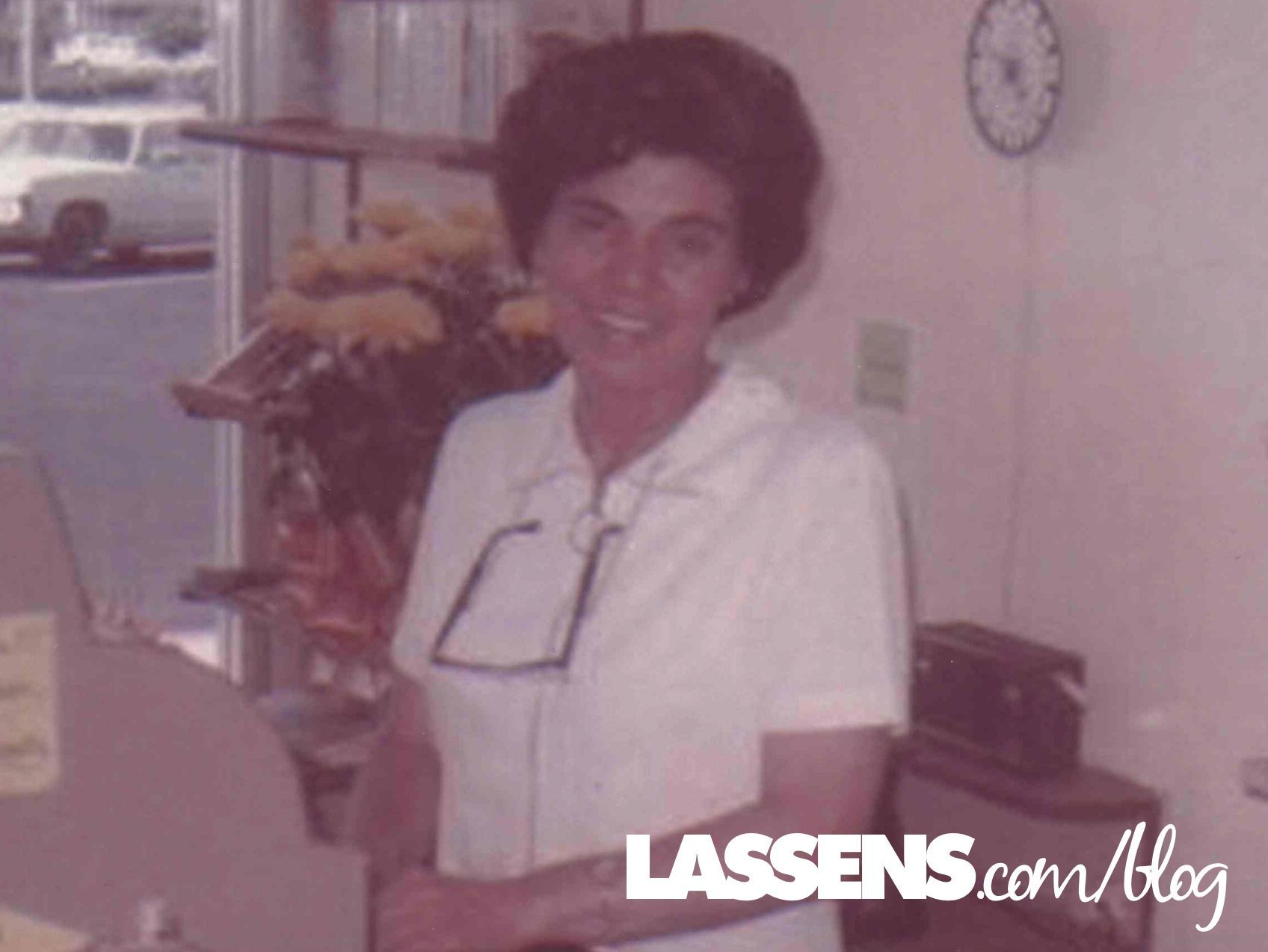 Woman in white shirt, smiling, holding eyeglasses, standing in a store with flowers and a clock.
