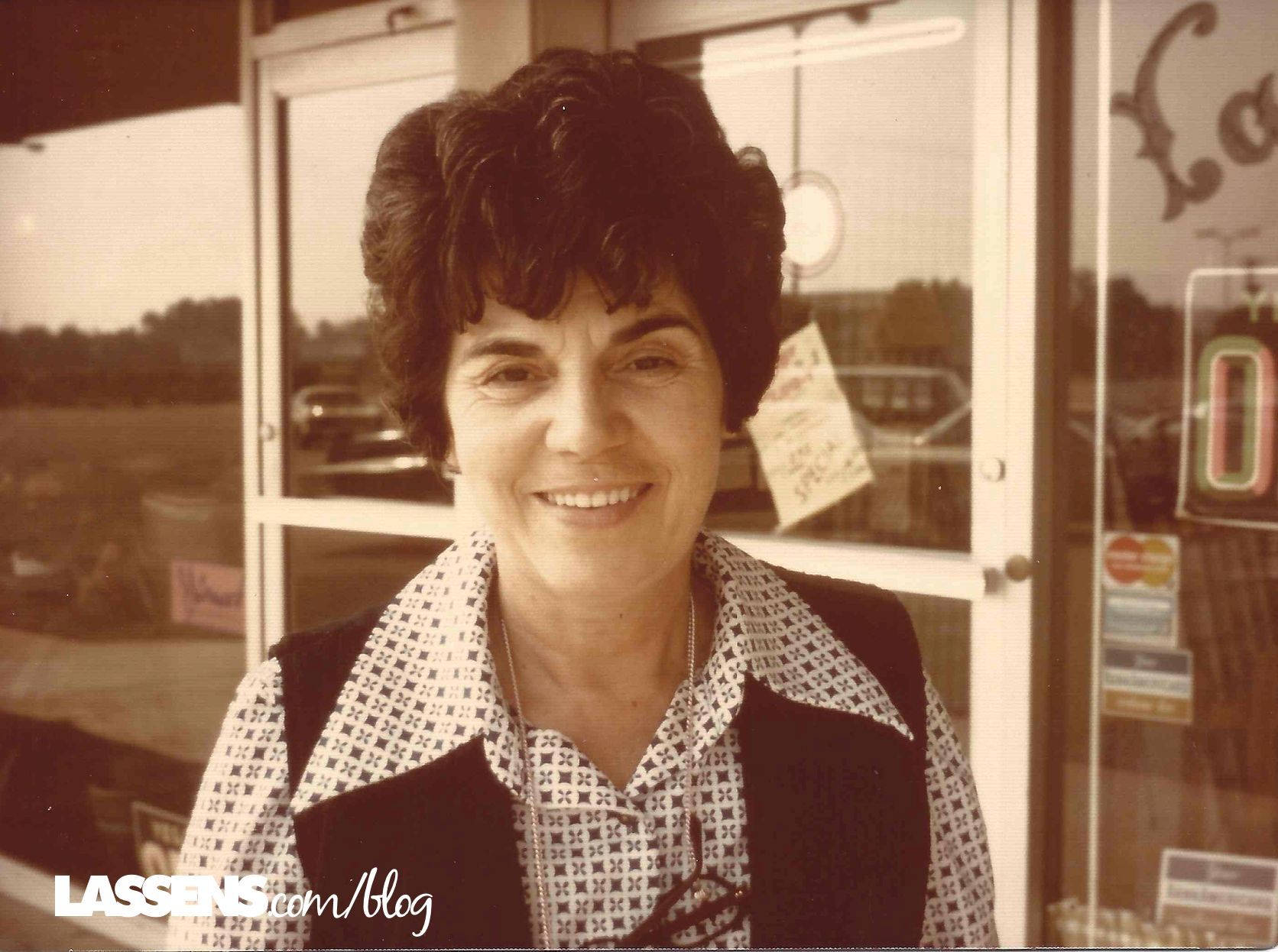 A smiling person wearing a dark vest over a patterned blouse, standing in front of a glass storefront.