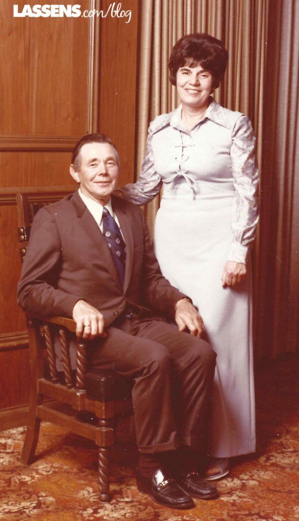 Man in a suit sits in a chair, woman in a white dress stands beside him. Studio portrait.