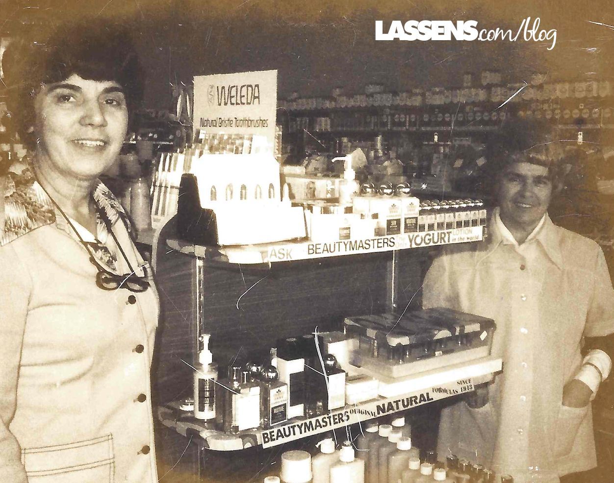 Two individuals stand smiling behind a store display counter filled with bottles and products, set in a retail space.