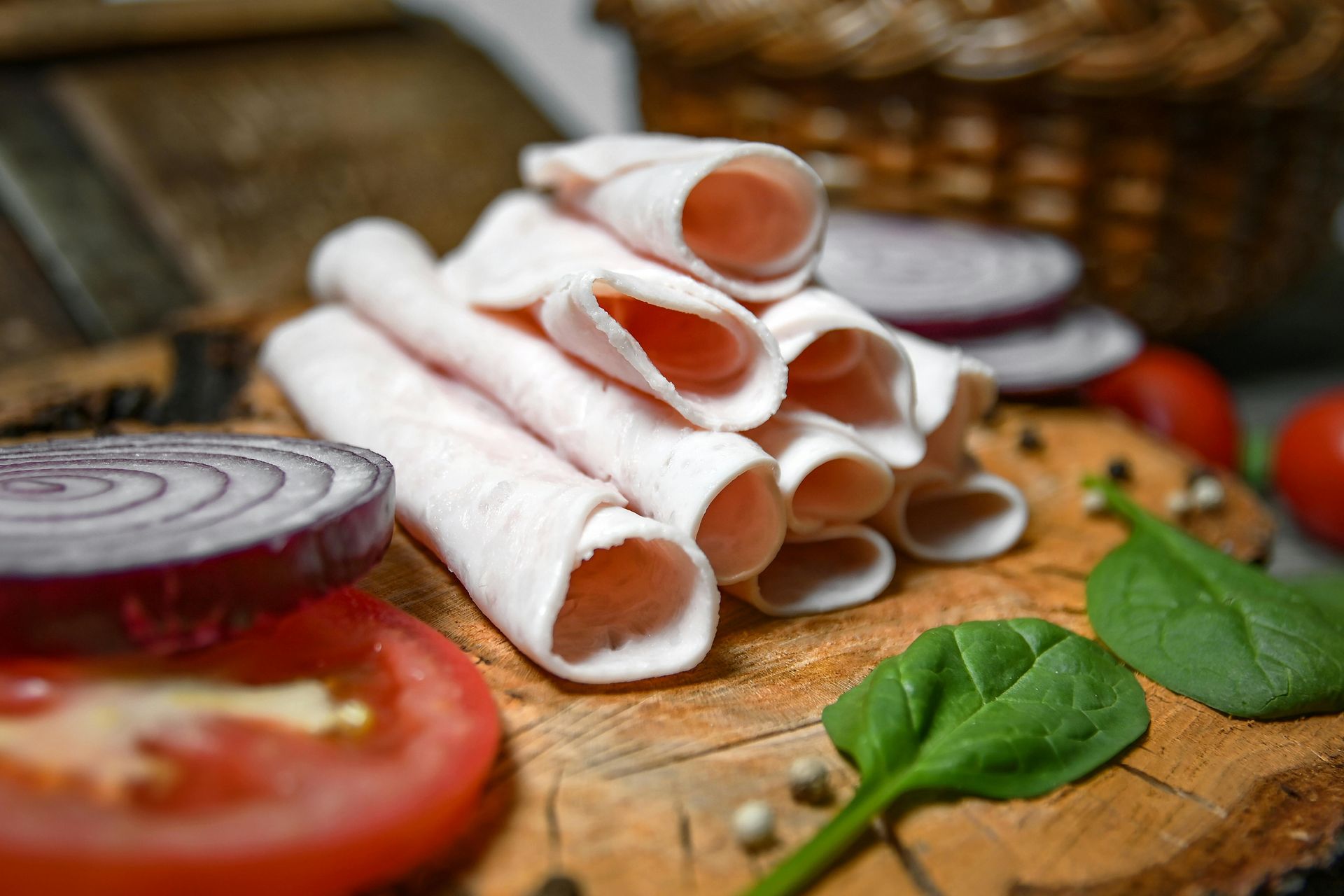 Rolled deli meat slices on a wooden board with onion rings, a tomato slice, spinach leaves, and peppercorns.