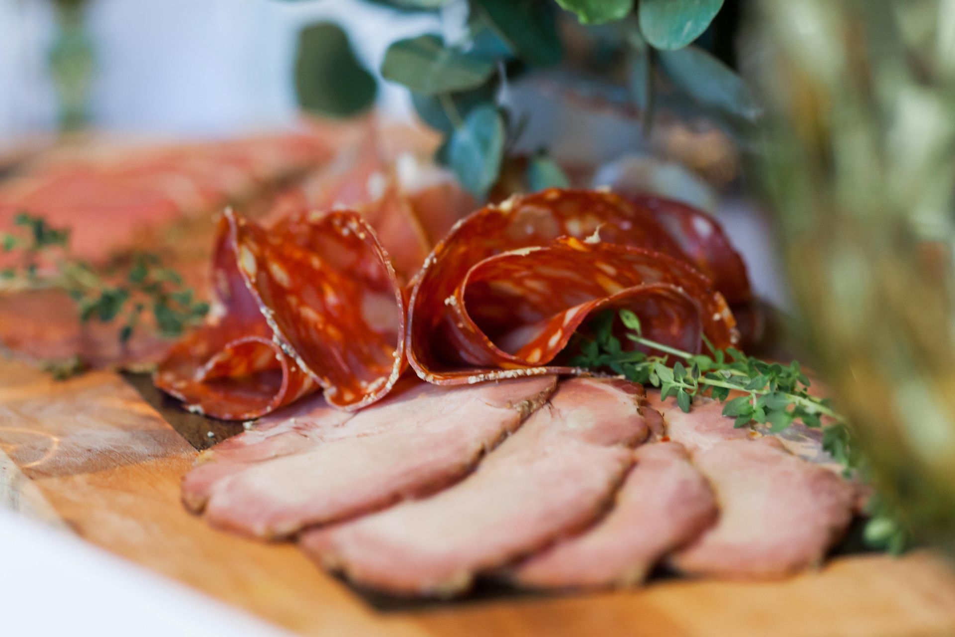 A wooden board holds neatly arranged slices of cured meat, including folded chorizo and roasted ham garnished with herbs.