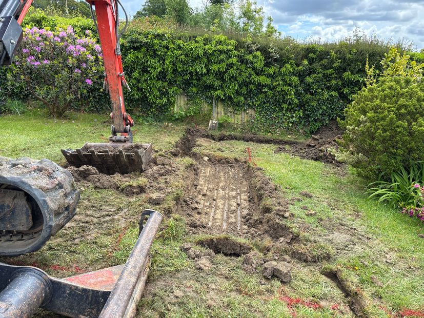 Excavator arm digging a patchy lawn beside hedges and flowers in a garden.
