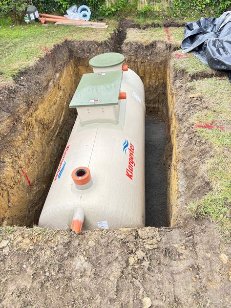 Workers installing a large silver underground tank at a suburban construction site with an excavator nearby
