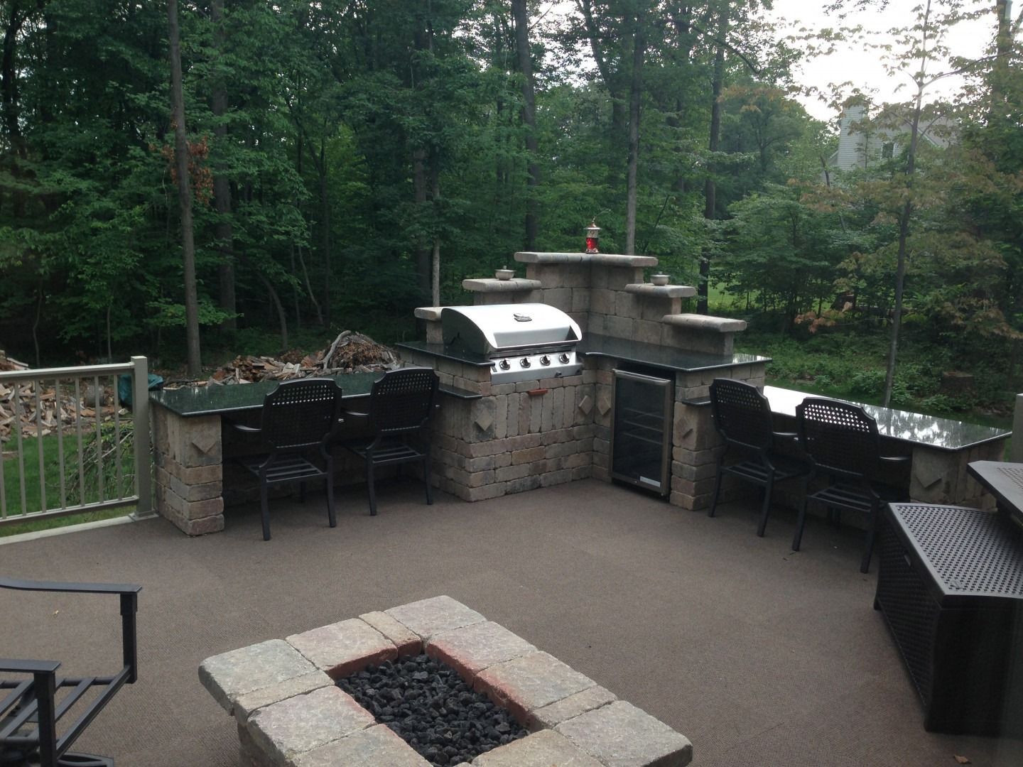 Stone patio with built-in grill and fire pit, surrounded by black chairs and overlooking wooded backyard.