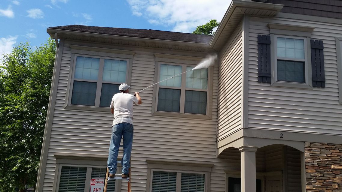 Person power washing a two-story beige house with a ladder on a sunny day.