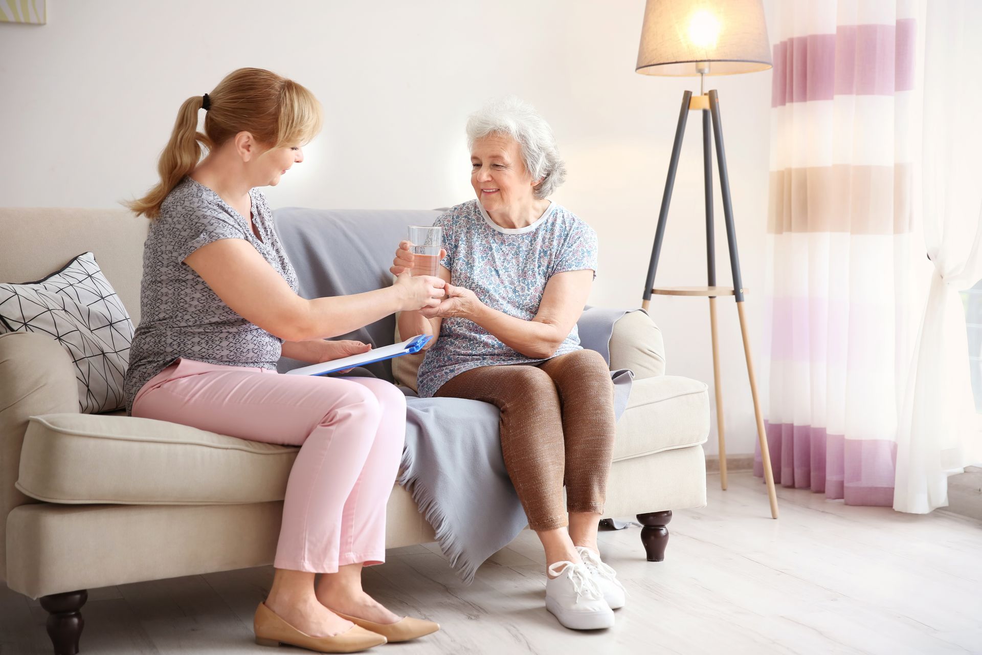Caregiver and older adult sitting on a sofa, sharing a cup in a bright living room.