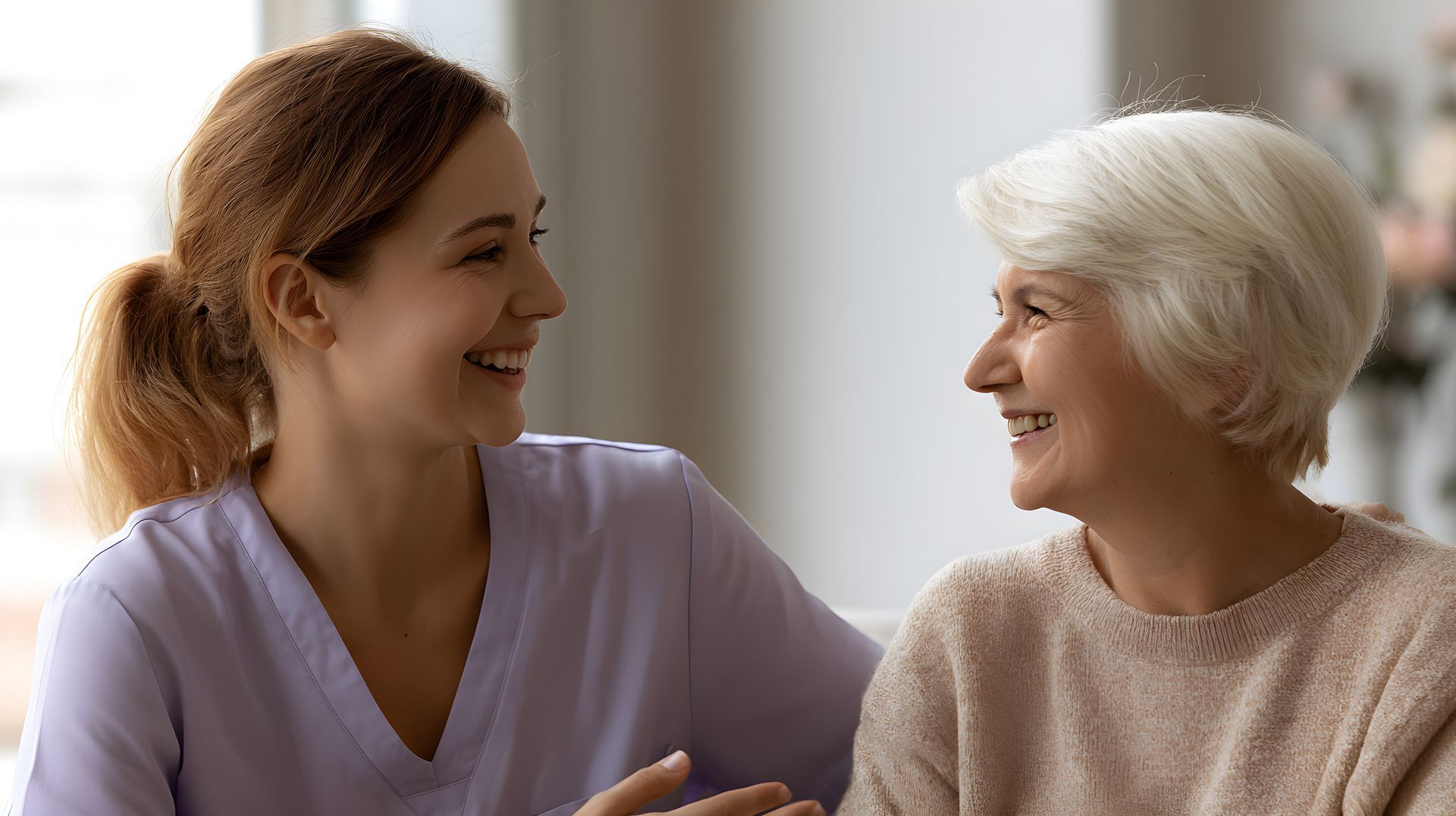Two women smiling at each other in a bright room, one in a lavender top and one in a beige sweater.