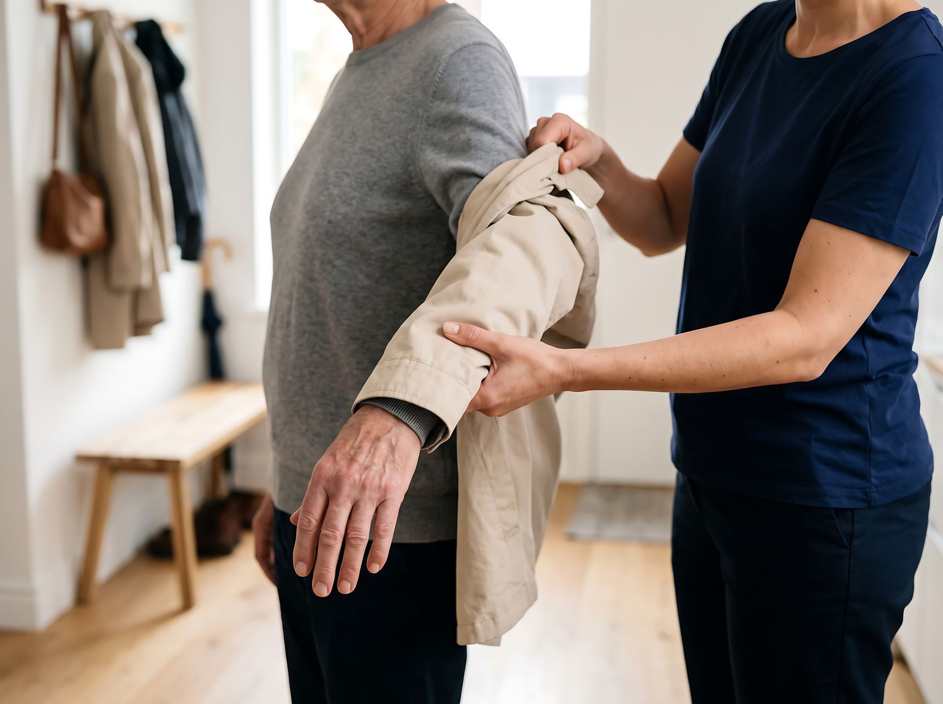 Caregiver helping a person put on a beige jacket in a bright room