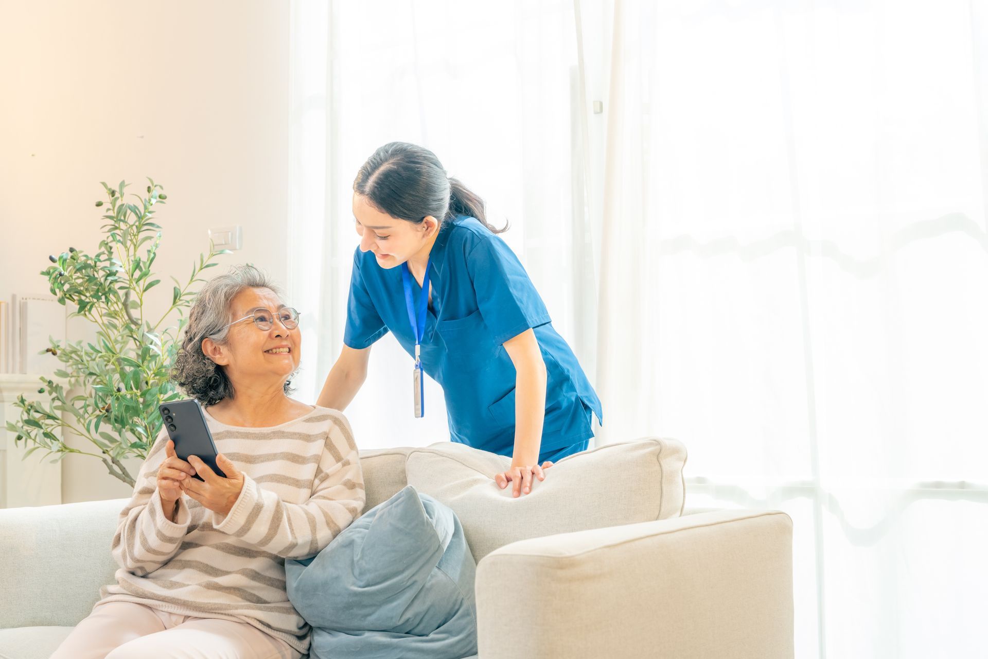 Caregiver in blue scrubs helping a smiling patient hold a smartphone in a bright living room