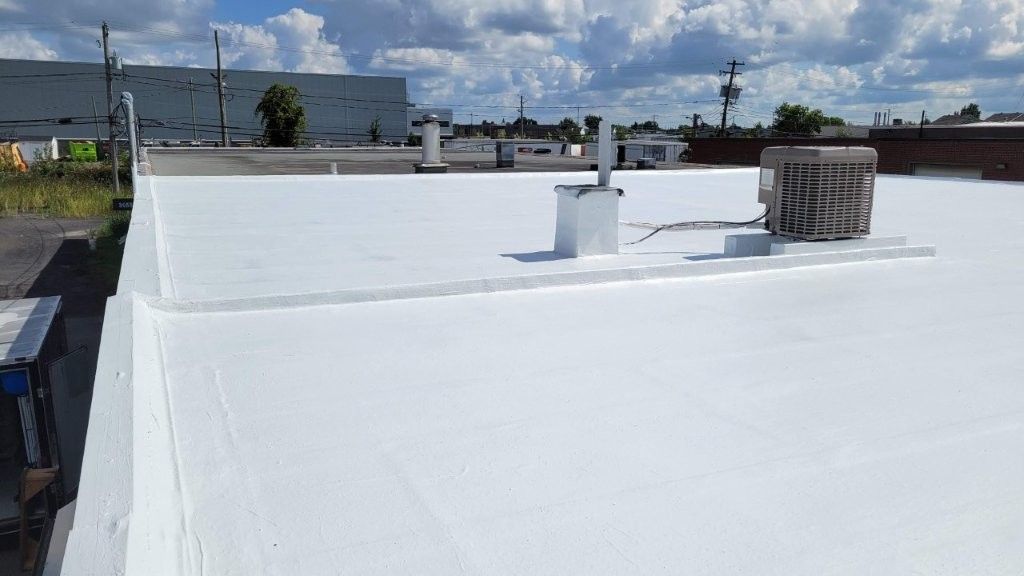 Toit plat blanc d'un bâtiment sous un ciel partiellement nuageux. On y voit des aérations et un climatiseur.