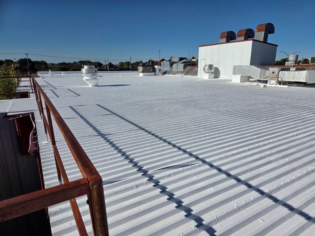 Toit commercial plat, recouvert d'une peinture blanche, avec une charpente métallique, des aérations et une rambarde rouillée se détachant sur un ciel bleu clair.