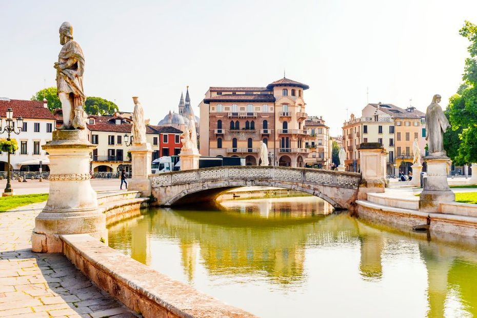 Fossato in Prato della Valle, Italia, fiancheggiato da statue, con edifici e la guglia di una chiesa sullo sfondo.