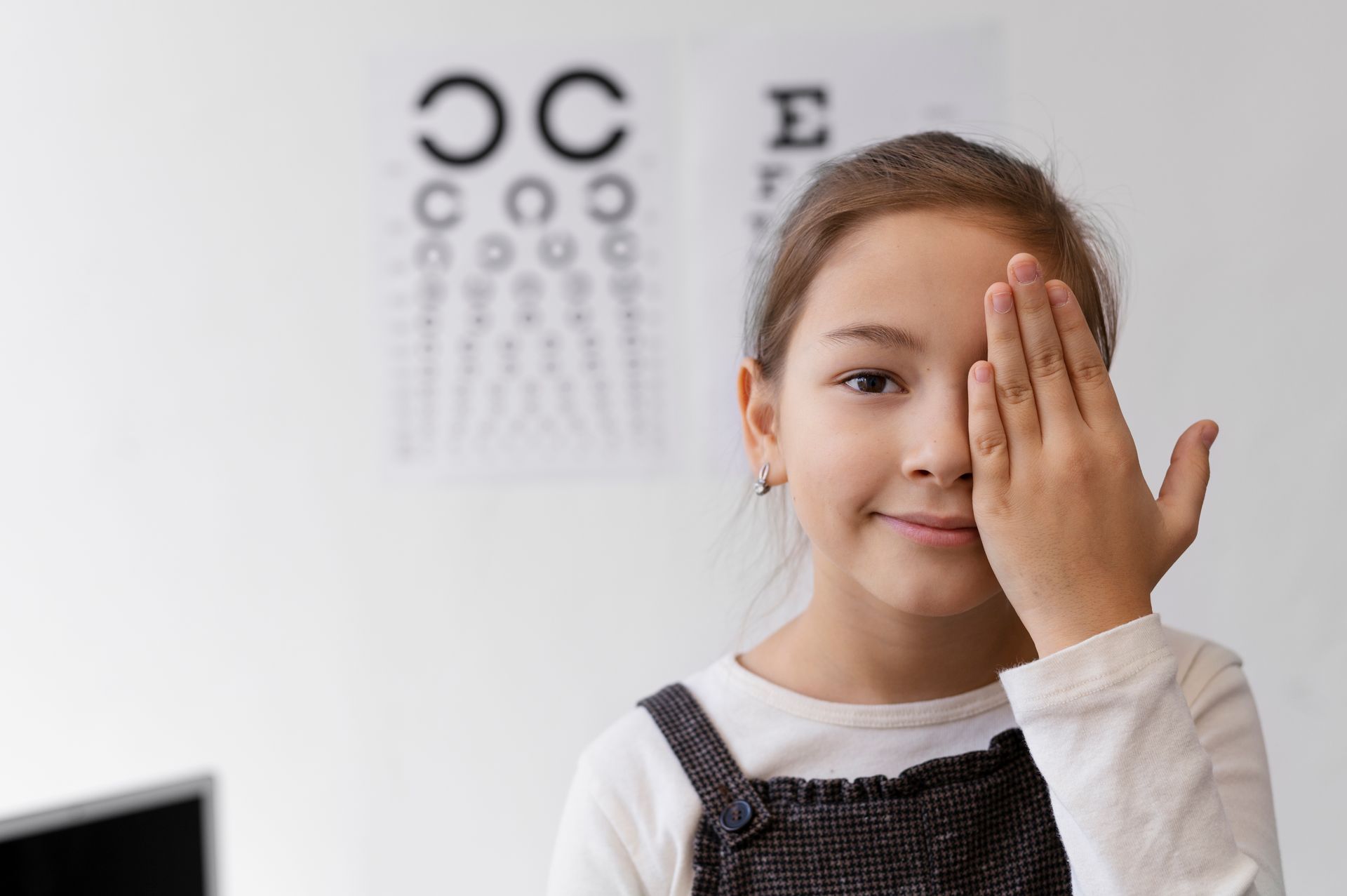 Young girl covering one eye with her hand during an eye exam, Snellen chart in the background.