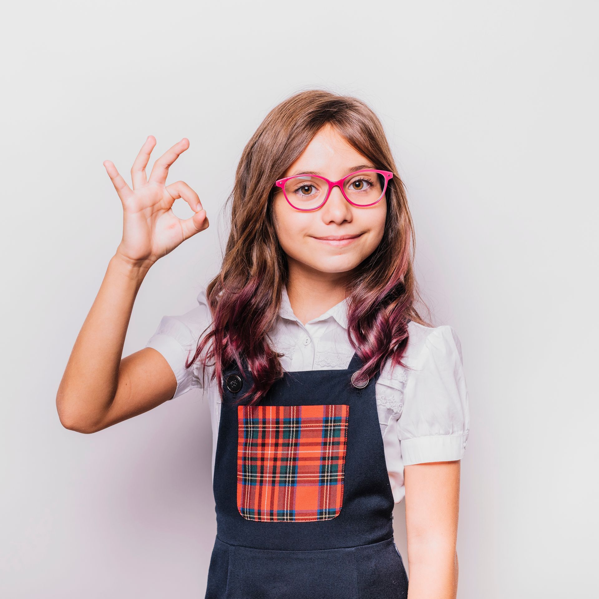 A young girl wearing glasses and overalls is giving an ok sign.