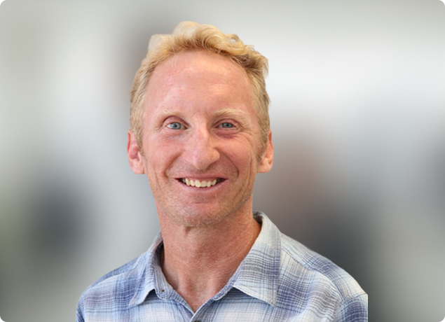 Smiling man in a light blue checkered shirt against a blurred gray background