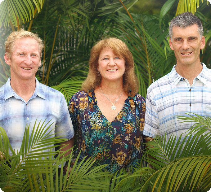 Three people standing in front of lush green tropical plants, smiling at the camera