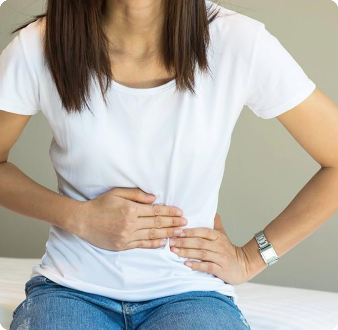 Person sitting with both hands on lower abdomen, wearing a white shirt and jeans, suggesting stomach pain