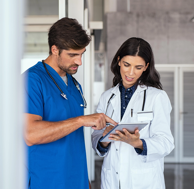 Two medical professionals in scrubs reviewing a tablet in a clinic hallway