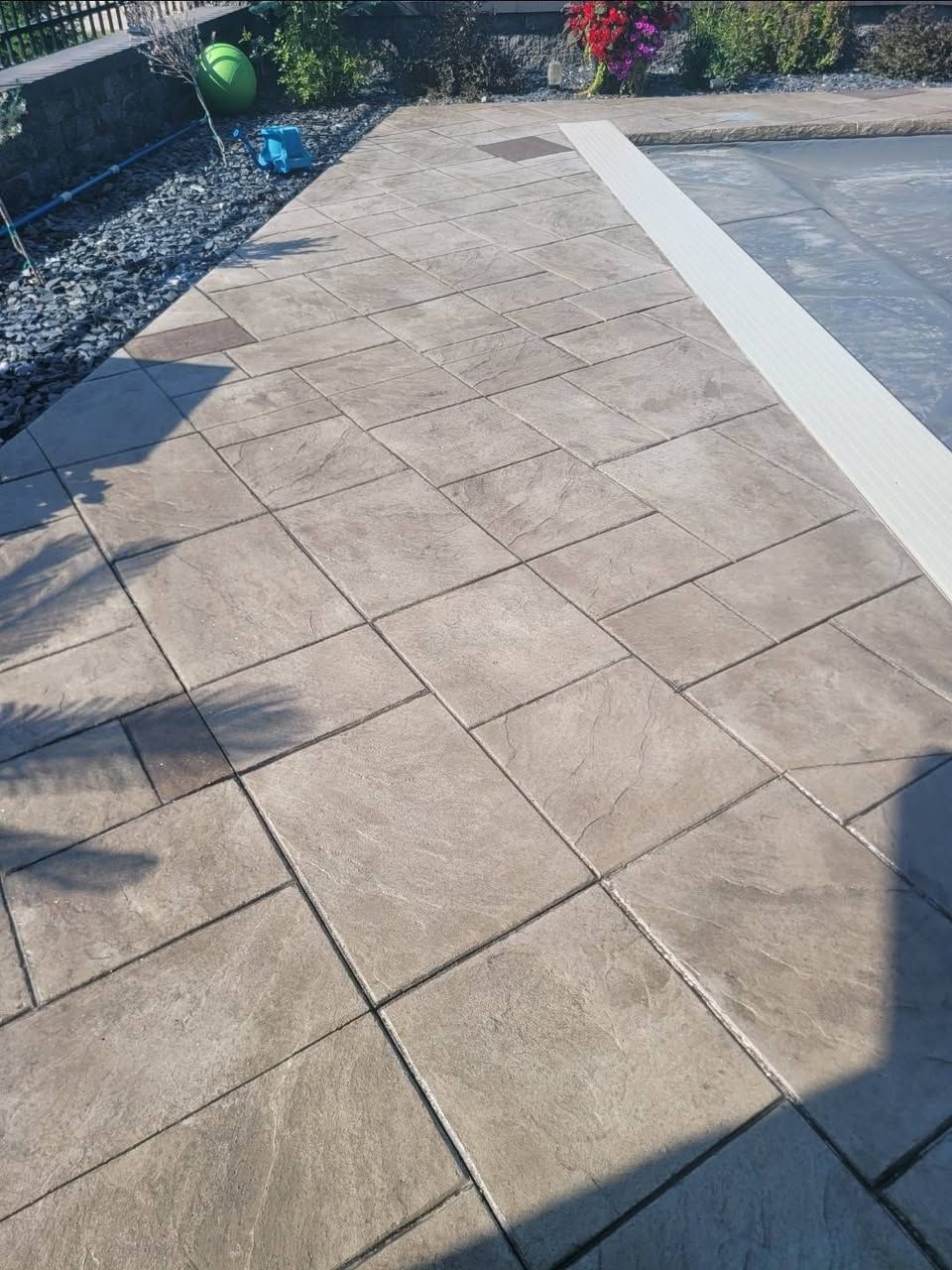 Sunlit beige stamped concrete stone patio with square pavers beside a gravel border and plants