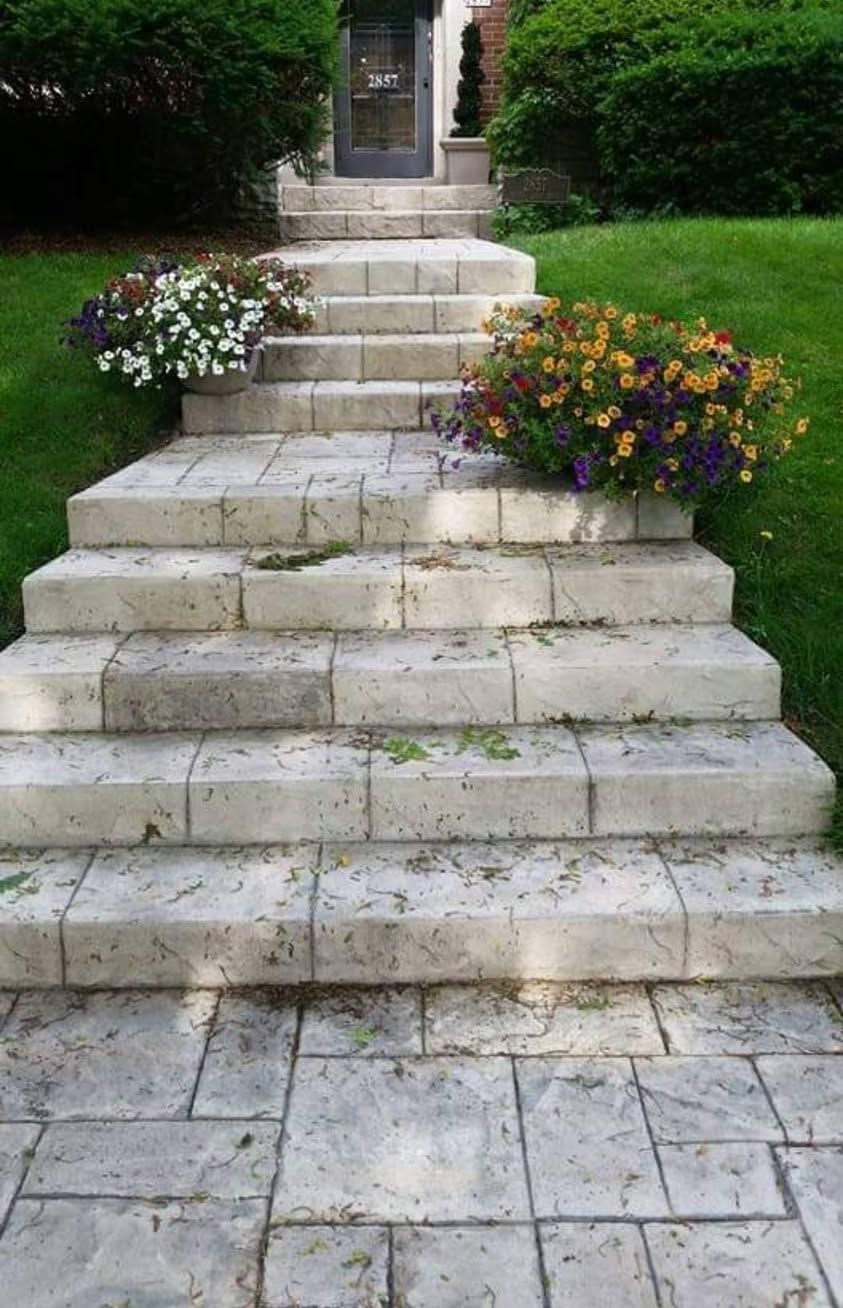 Stamped stone steps in a garden leading to a doorway, flanked by white and purple flower planters.