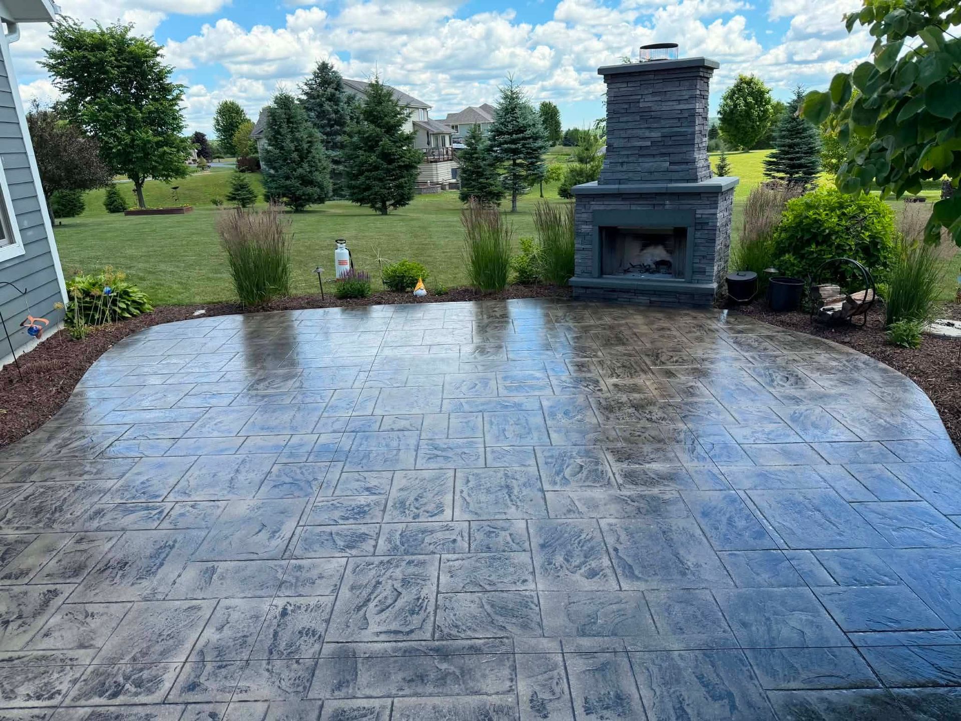 Wet stamped-concrete patio with stone fireplace, framed by shrubs and a grassy backyard under a cloudy sky