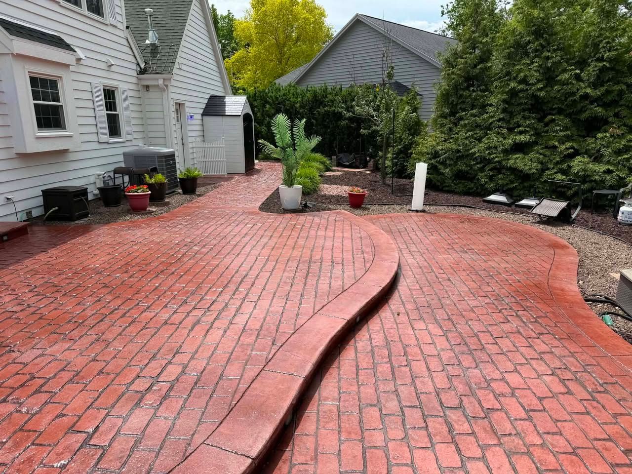Red brick stamped concrete patio with a raised curved strip, potted plants, shrubs, and a white house beside a driveway